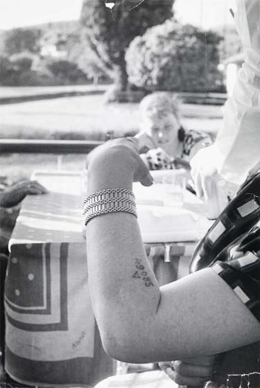 Black and white photograph: The photo shows the bent arm of a woman. The number “49085” is tattooed on her forearm. Below it is a triangle. She is wearing a bracelet on her wrist. A table, a woman, trees and a river can be seen blurred behind her.