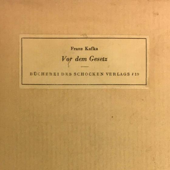 Ausschnitt aus einem gelblich-beigen Buchcover mit aufgeklebtem Etikett auf dem steht: „Franz Kafka, Vor dem Gesetz, Bücherei des Schocken Verlags / 19“.