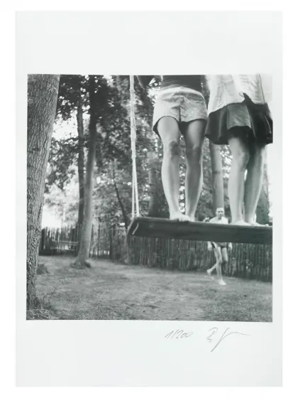 A black and white photo of two girls standing on a wide wooden swing. Only their legs are visible