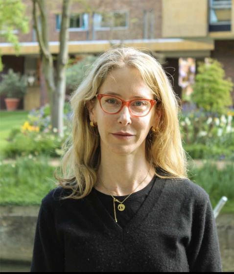 Porträtfoto einer Frau mit langen blonden Haare, roter Brille und schwarzen Pullover, im Hintergrund ein kleiner Stadtgarten und eine Hausfassade.