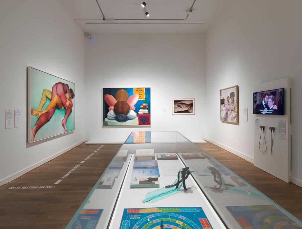 Exhibition view, a glass display case stands in the middle of the room, colorful pictures hang on the walls.