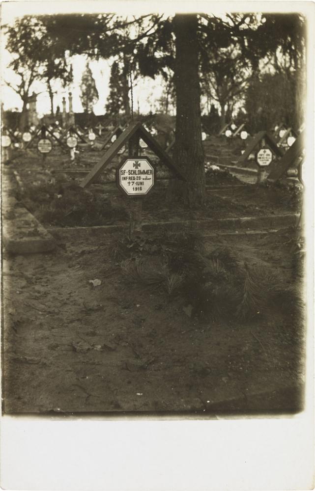 Black-and-white photograph: gravestone with cross and inscription; more graves in the background