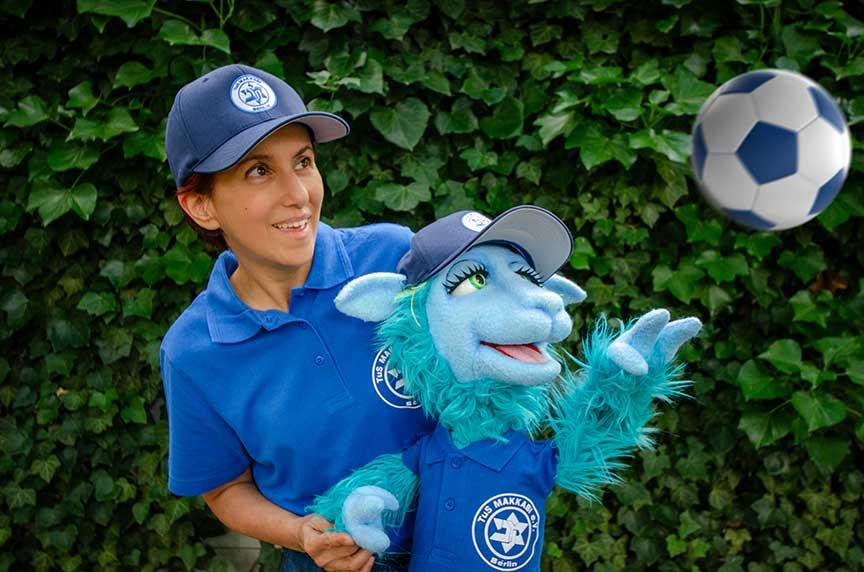 Woman with hand puppet in front of a hedge. Both are wearing Tus Maccabi Berlin shirts and caps and are watching a soccer.