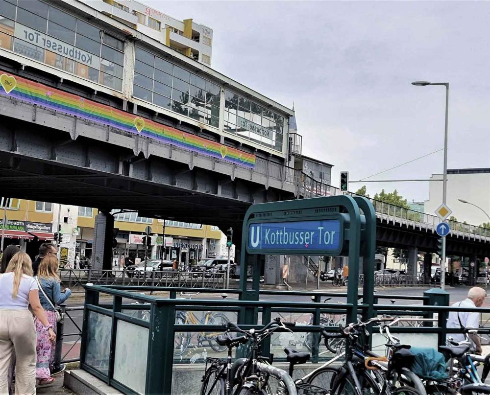 Straßenszene am Kottbusser Tor in Berlin: Eingang zur U-Bahn mit parkenden Fahrrädern, Fußgänger, die Hochbahntrasse und Hochhäuser im Hintergrund.