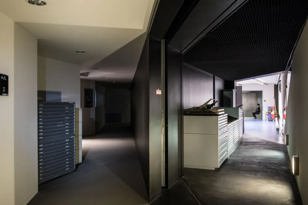A security guard is sitting on a chair at the far end of a corridor to the right. To the left there is another corridor, unlit.