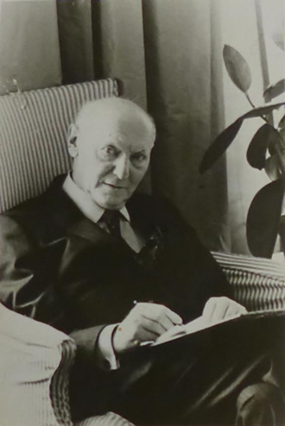 B-W photo of Isaac Bashevis Singer. He is sitting in an armchair, writing and looking into the camera.