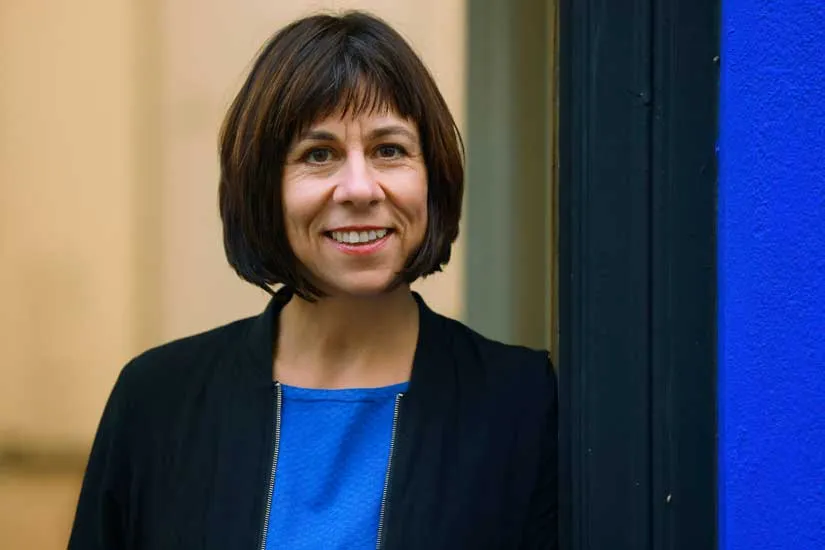 Upper body and head of a woman smiling at the camera. She is wearing a blue top and a black blazer over it.