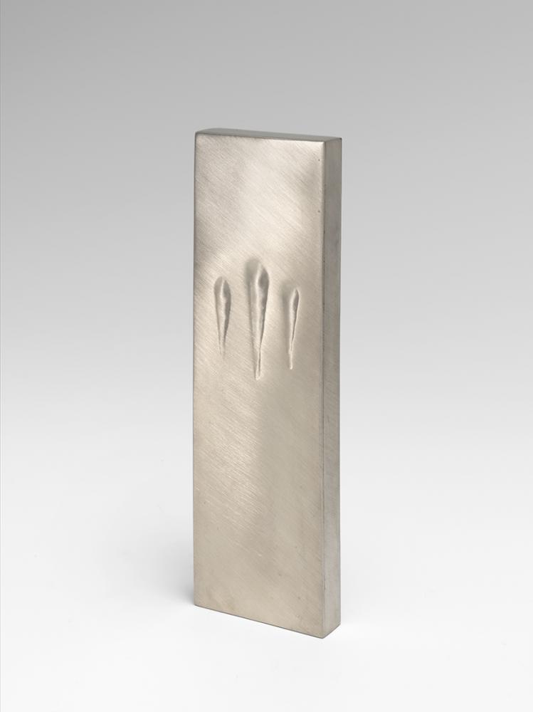 The image shows the art work Touching Mezuzah by Sari Srulovitch. The object ha s a rectangular shape and is made of silver. 