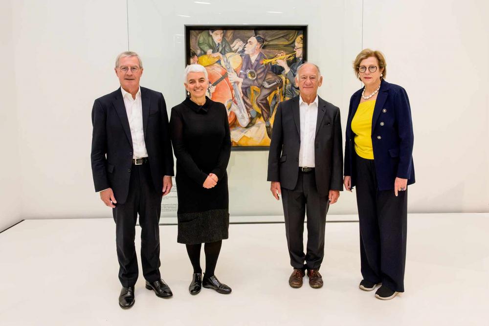 Four people are standing in front of a painting of a jazz band.