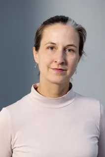 A woman is shown with her face and upper body, wearing a pink sweater and looking directly into the camera.
