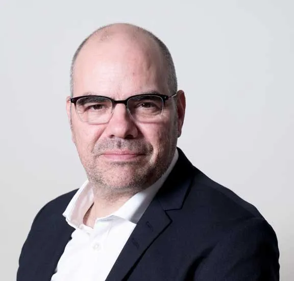 A man with a receding hairline and glasses looks directly into the camera. He is wearing a white shirt and a black jacket.