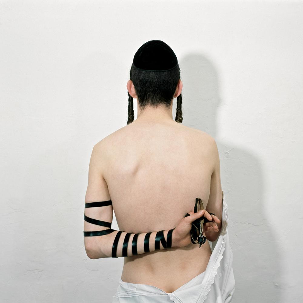 The iamge shows the art work Tefillin schel Jad by Benyamin Reich. The photography shows the naked back of a young man wearing a kippa and the traditional leather straps wrapped in a unothodox manner.