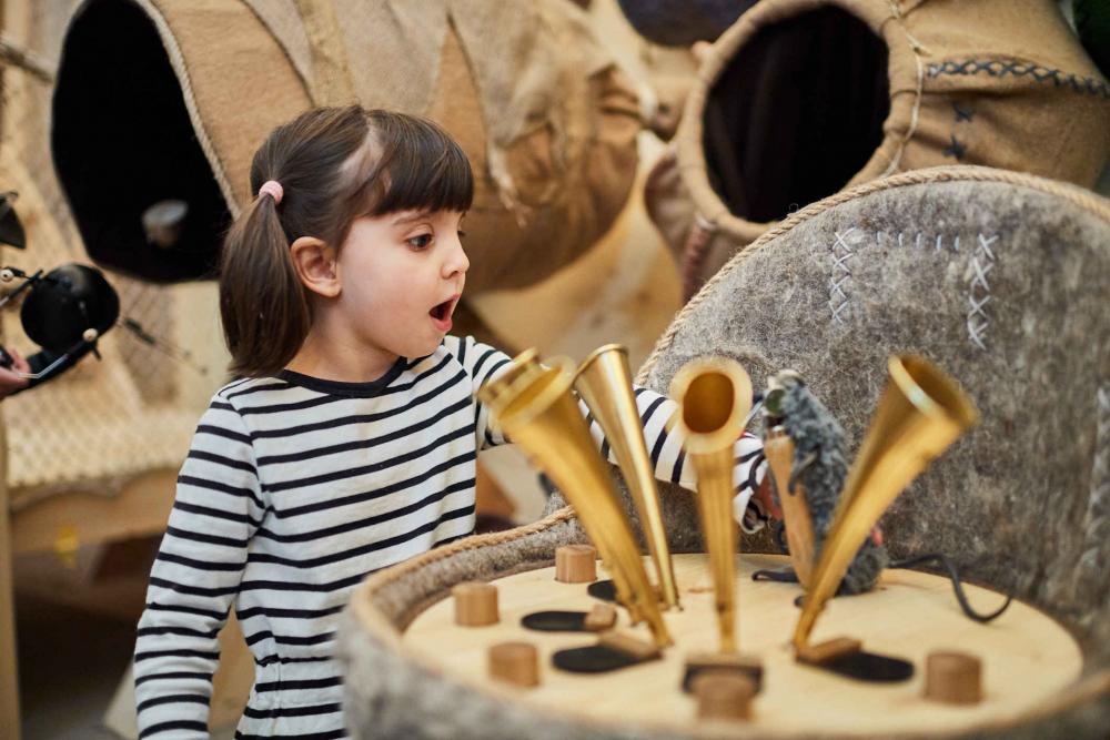 Child wearing a striped sweater stands in front of funnels standing on a wooden element and has opened his mouth