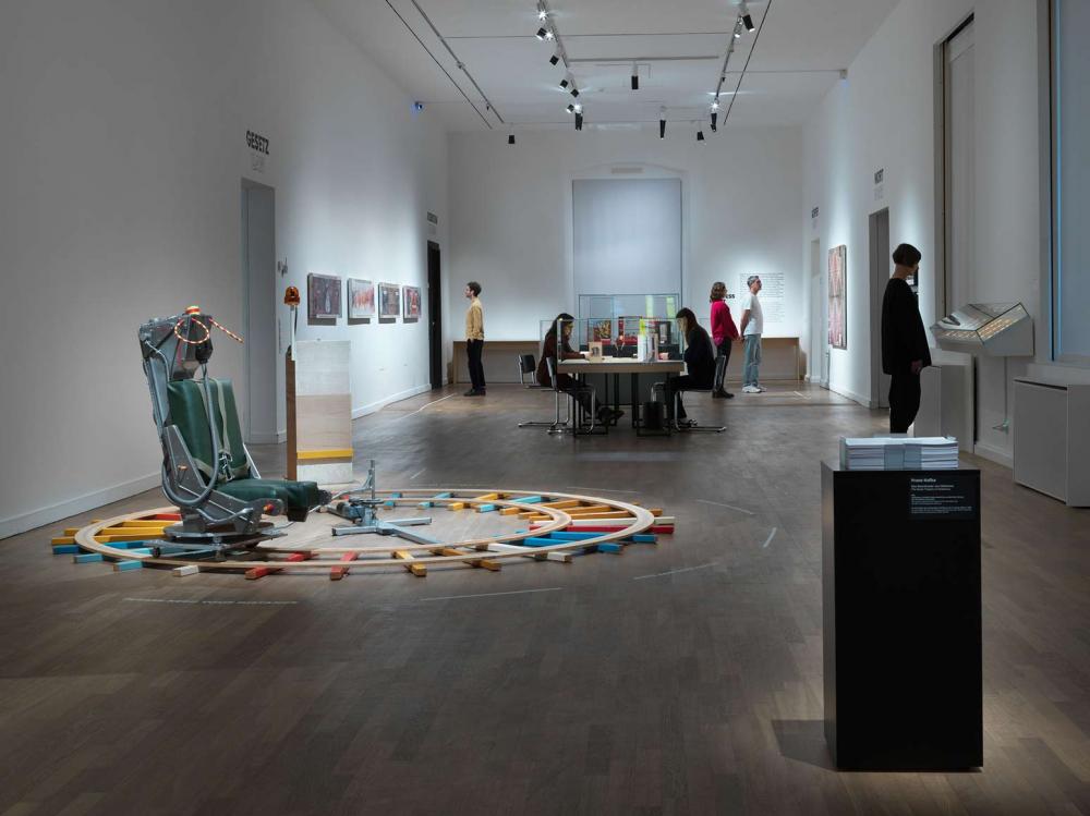 View into an exhibition space with visitors, in the center of the room an installation with an ejection seat on wooden rails running in a circle.