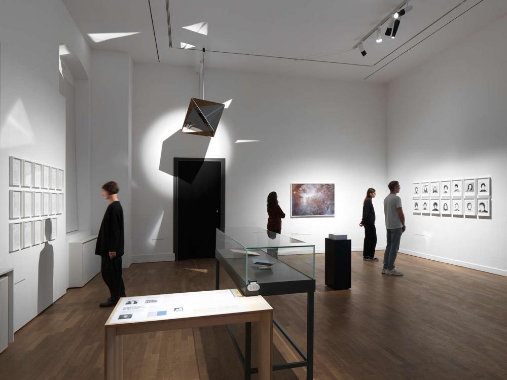 View of an exhibition room with visitors looking at paintings and photos on the walls.