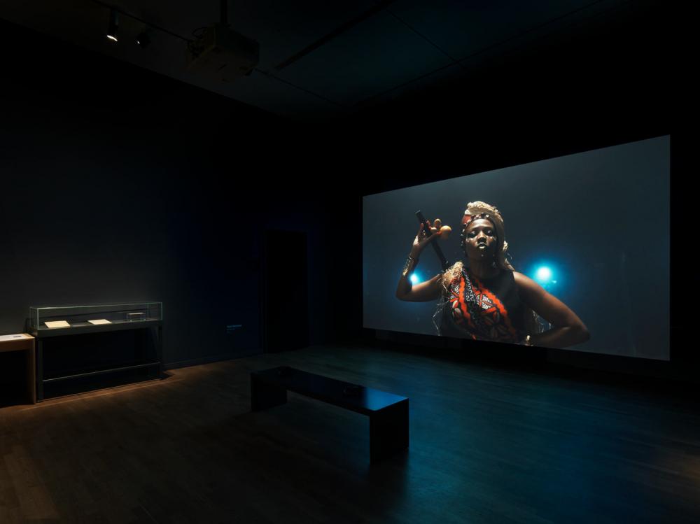 View into an exhibition room with a movie screen, on it a female and Caribbean read person with red and black sash, headdress made of straw and staff with calabash in her hand in front of a dark background.