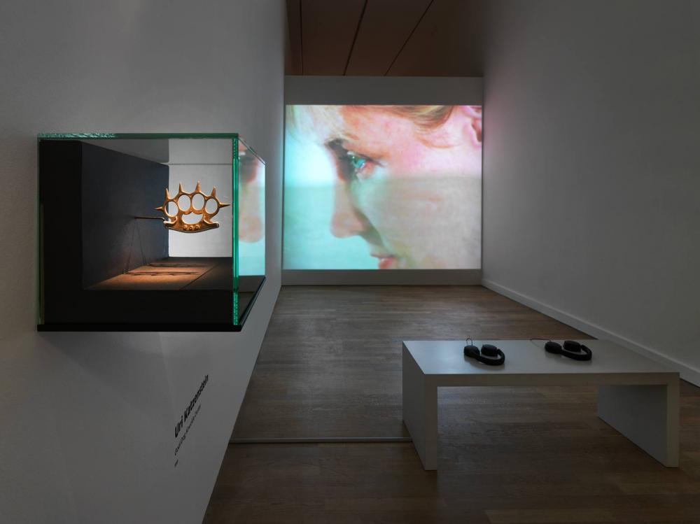 View into an exhibition room, in the foreground a display case in which a knuckleduster is exhibited, in the background a screen on which a close-up of a face can be seen, in the middle of the room a bench with two headphones.