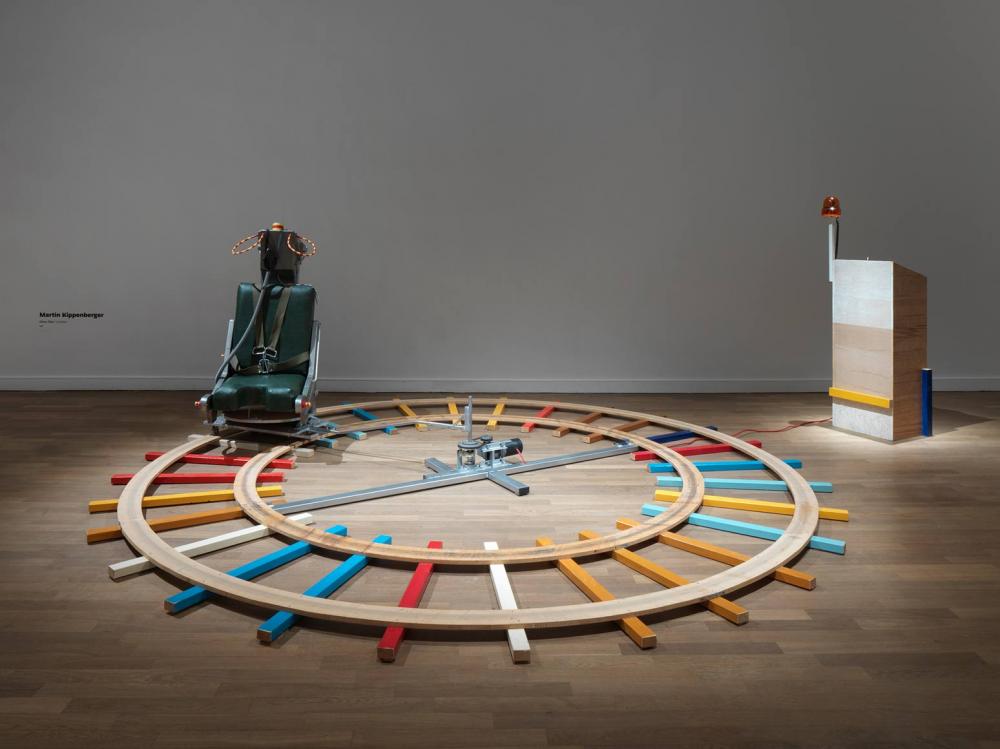 Ausstellungsraum mit im Kreis laufenden bunten Holzschienen auf dem Boden, darauf ein Schleudersitz und rechts daneben ein Holzpult mit orangenem Licht, das mit dem Sitz verkabelt ist.