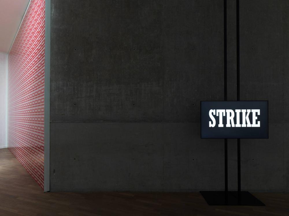 View into a exhibition room with a gray concrete wall, in front of which is a sign with bright white letters on a dark background that reads: STRIKE.