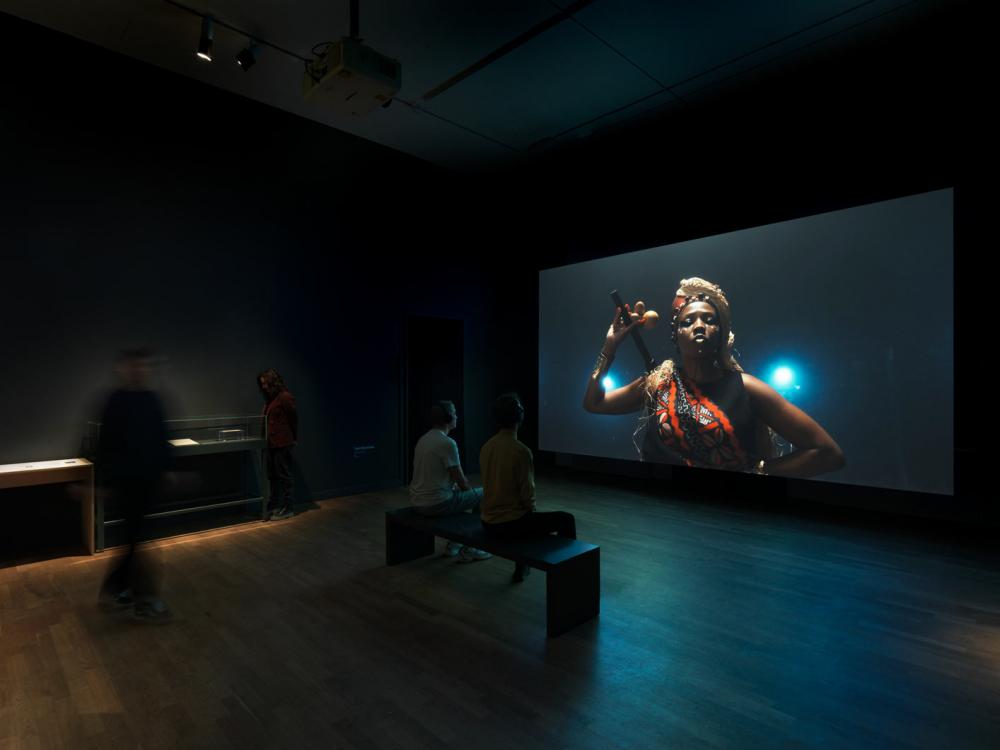 View into an exhibition room with a movie screen, on it a female and Caribbean read person with red and black sash, headdress made of straw and stick with calabash in hand in front of a dark background.