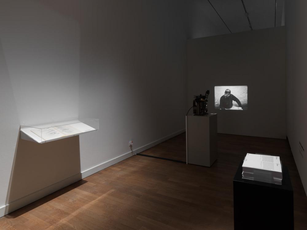 View into a darkened exhibition room with a black-and-white film projection on the wall, in the foreground a stack of papers on a black pillar.