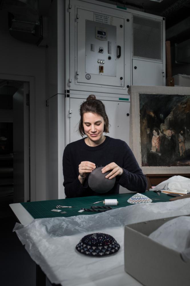 Eine junge Frau sitzt an einem Arbeitstisch in einem Atelier und bestickt Kippas.