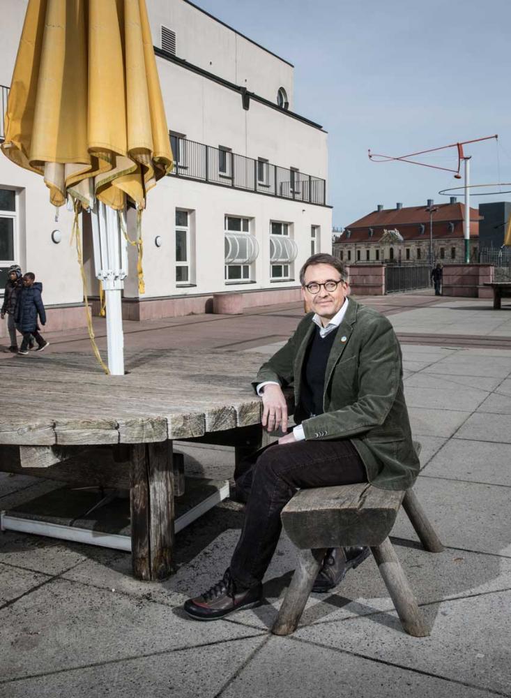 Ein Mann mit Brille und grünem Sakko sitzt an einem Holztisch mit gelbem Sonnenschirm auf einer Dachterasse. Im Hintergrund ist das Jüdische Museum Berlin zu sehen.