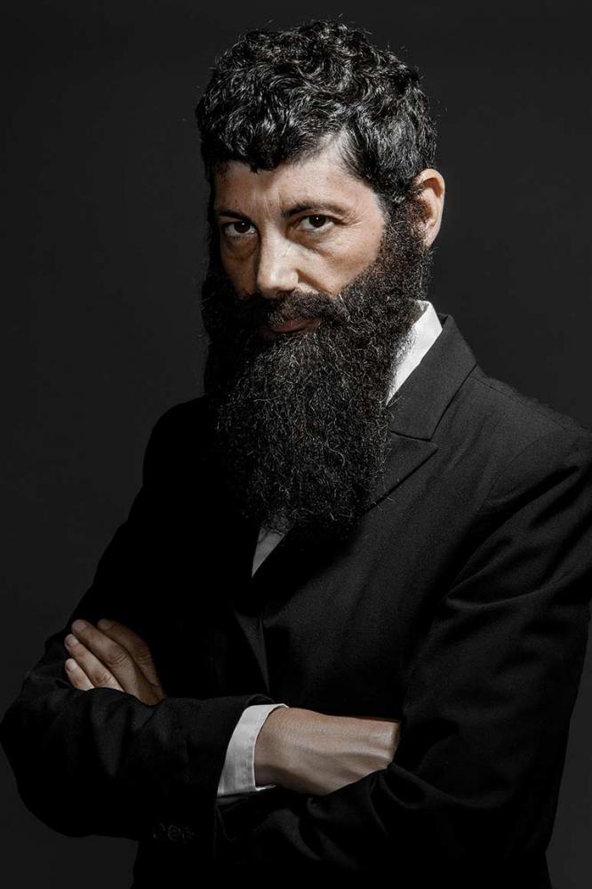 Half-length portrait of a bearded man with long, dark full beard and dark hair. He wears a suit and crosses his arms in front of his chest. The left half of the body is facing the viewer, the face can be seen in half profile. 