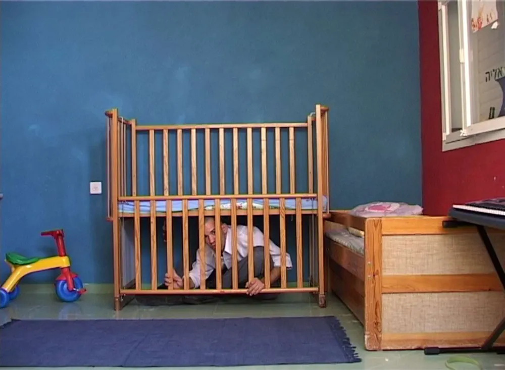 An adult sits on the floor of a child's room under the crib for babies and looks through the lowered wooden bars.