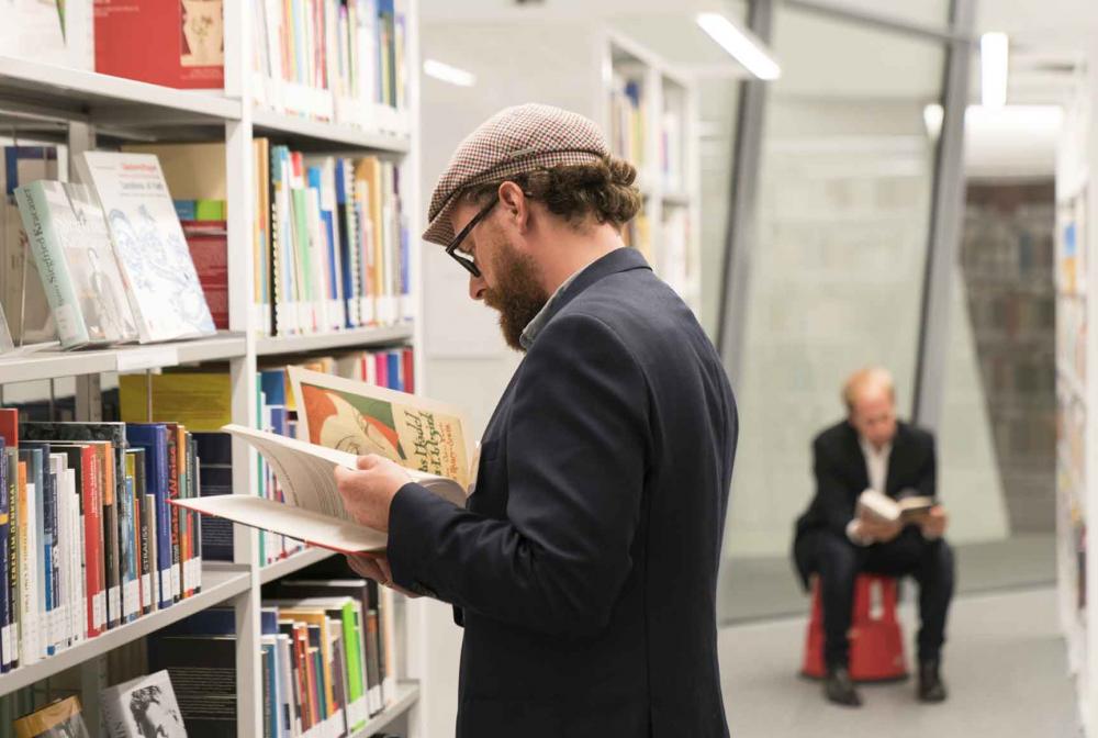 A man in a suit, glasses and cap stands in front of a bookshelf and leafs through a book. In the background, another man is sitting on a red stool with a book in his hand.