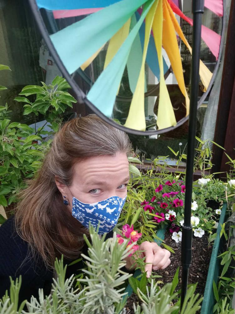 Selfie von Mirjam Bitter mit blauer Maske, die von Pflanzen umgeben auf ihrem Balkon steht – im Vordergrund ein regenbogenfarbenes Windrad.