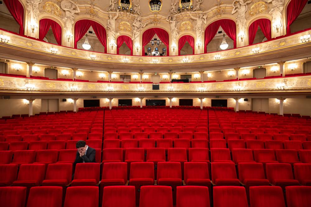 Fotografie einer Person allein auf einem der roten Sitze im Parkett eines leeren Theaters, den Kopf in die Hände gestützt