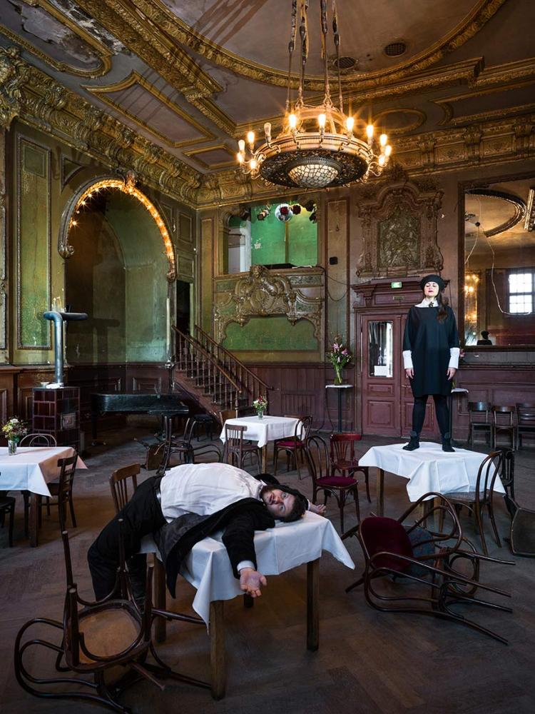 Two people in a room with tables and chairs, a grand piano and golden stucco on the ceiling: the woman is standing on one of the tables with white tablecloth, the man is lying with his back on another one, some chairs have fallen over