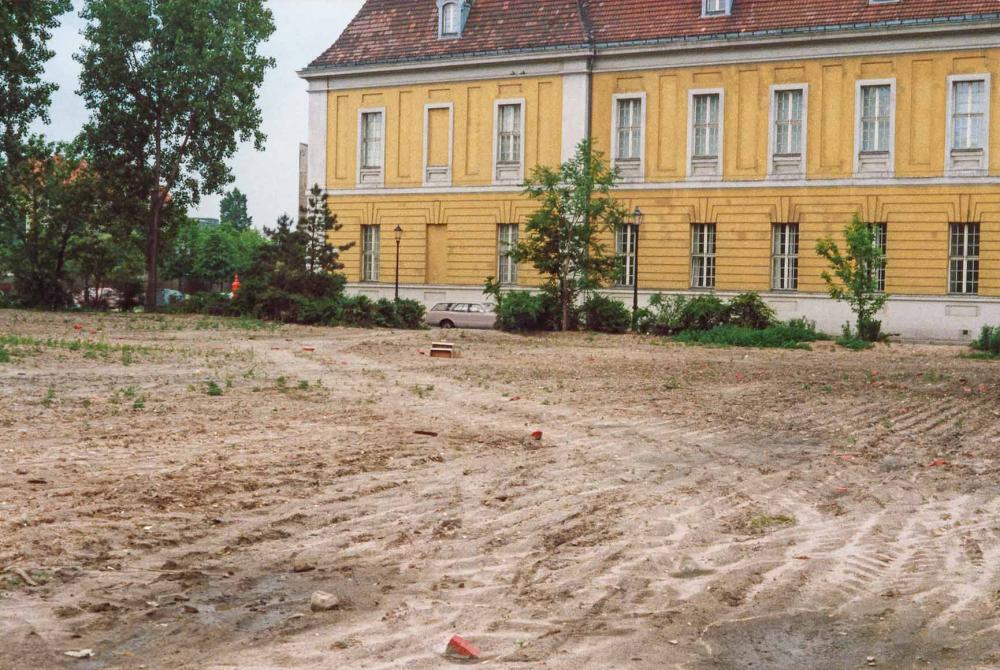 Sandy wasteland with scattered red-painted stones, in the background a wide building with a yellow façade and red roof.