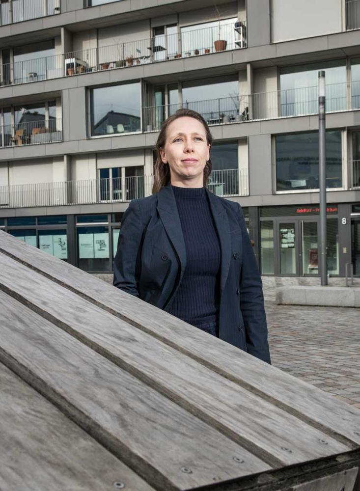 Eine Frau mit offenen braunen Haaren und einem dunkelblauen Blazer steht im Freien vor einem modernen Wohnhaus. 