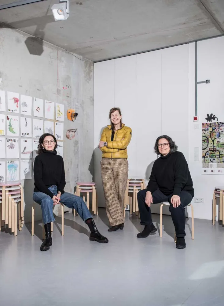 In a room with a concrete wall and ceiling, two women in black sweaters are sitting on wooden stools to the right and left. A third woman dressed in yellow is standing in the middle.