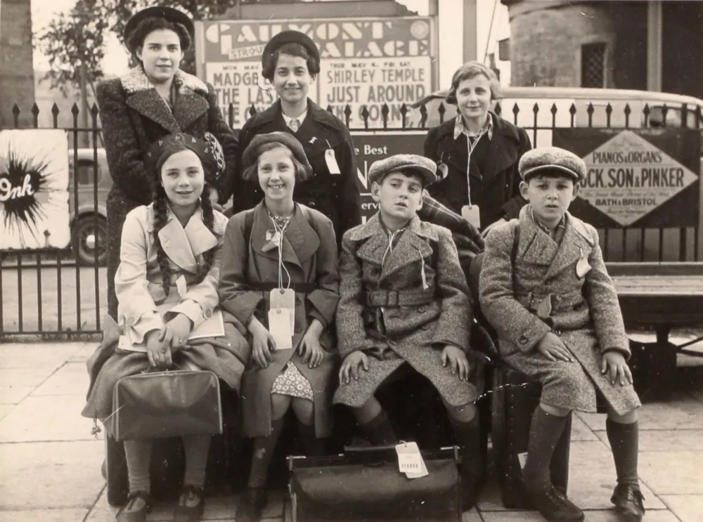 Black-and-white group photo of seven children in coats with baggage.