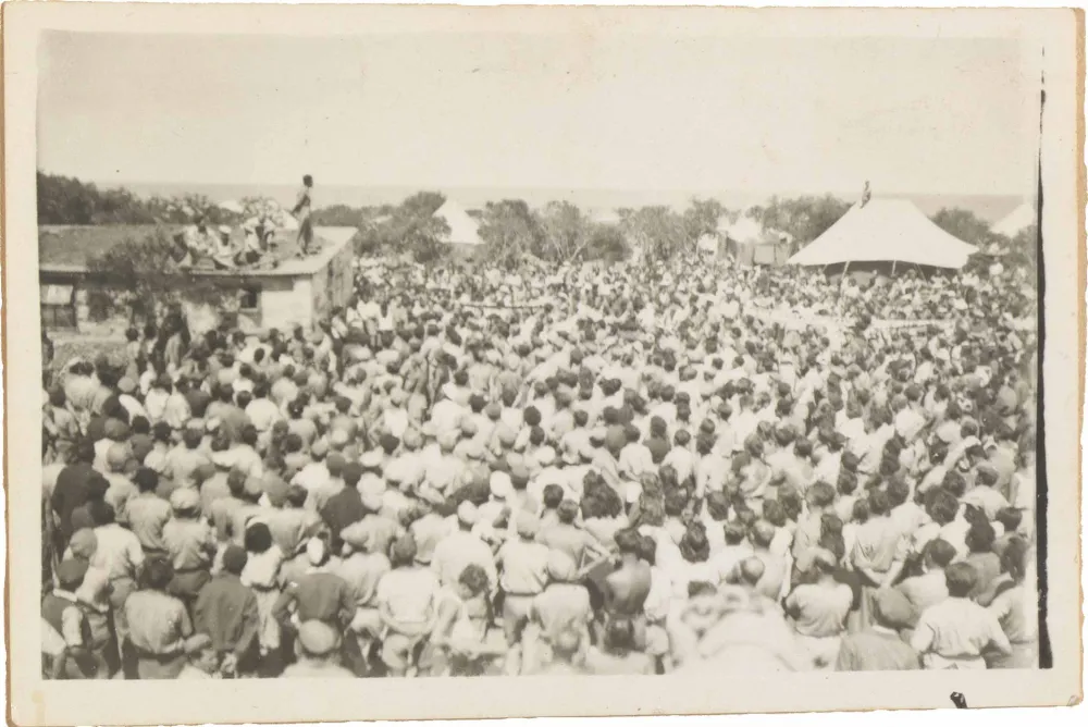 In the black-and-white photo taken from diagonally above, a large crowd of people is facing a house on whose roof a man is standing and speaking to the crowd.