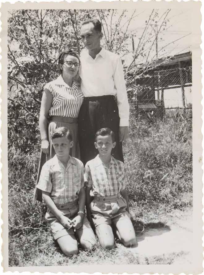  The photo shows a man, a woman and two boys outdoors. The children kneel on the floor. On the left behind them is the woman, on the right next to her is the man. In the background there is a garden and a building.