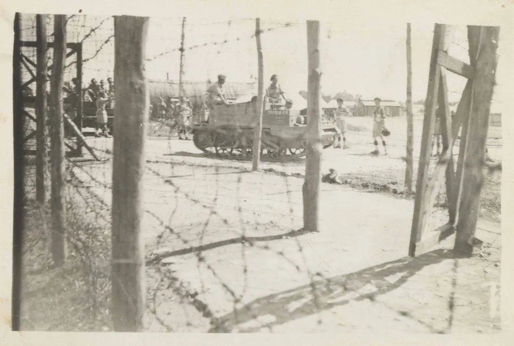 A barbed wire fence attached to wooden pillars can be seen in the foreground of the black and white photo. In the background are two tanks with soldiers sitting on them and a few internees standing around them.
