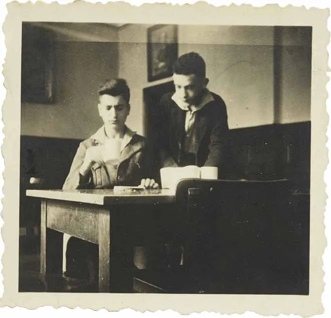Two boys at a table, one sitting with a cup, one standing diagonally behind him (black-and-white photo).