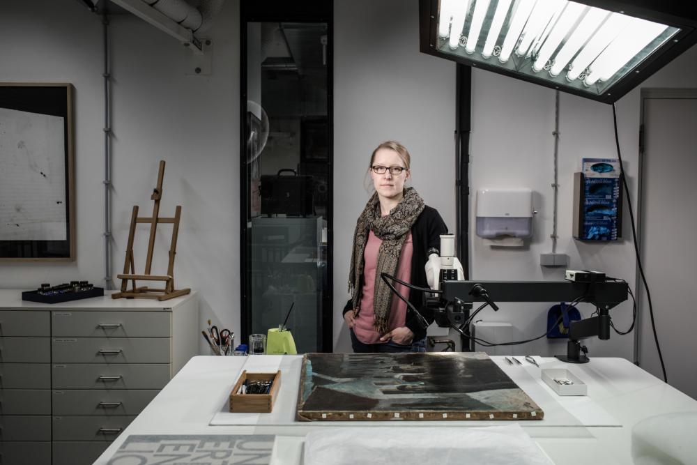 Eine junge Frau steht an einem Arbeitstisch in einem Atelier, vor ihr liegt ein altes Ölgemälde.