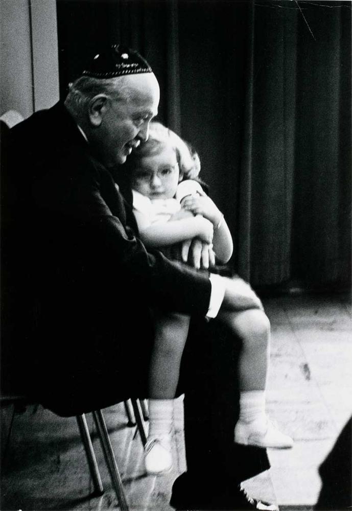 Black and white photograph: An elderly man in a dark suit wearing a kippah is sitting on a chair. He can be seen in profile hugging a little girl with glasses in light-colored clothing who is sitting on his lap.