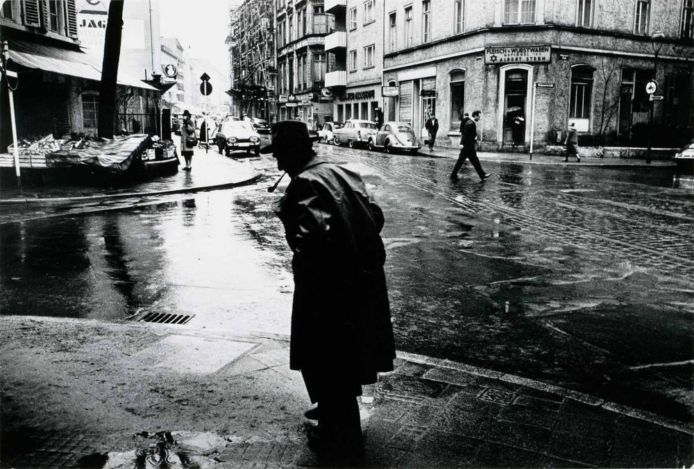 Schwarzweiß-Fotografie: Das Foto zeigt zwei Eckhäuser an einer regennassen Straßenkreuzung. Vor dem linken Eckhaus stehen mit Planen abgedeckte Verkaufstische. Das rechte Eckhaus beherbergt eine koschere Metzgerei, an der Fassade ist über dem Ladeneingang ein Schild angebracht, das die Aufschrift „Fleisch u. Wurstwaren Geflügel Albert Stern“ trägt. Im Bildvordergrund ist ein älterer Mann mit Mantel, Hut und Pfeife im Mund zu sehen.