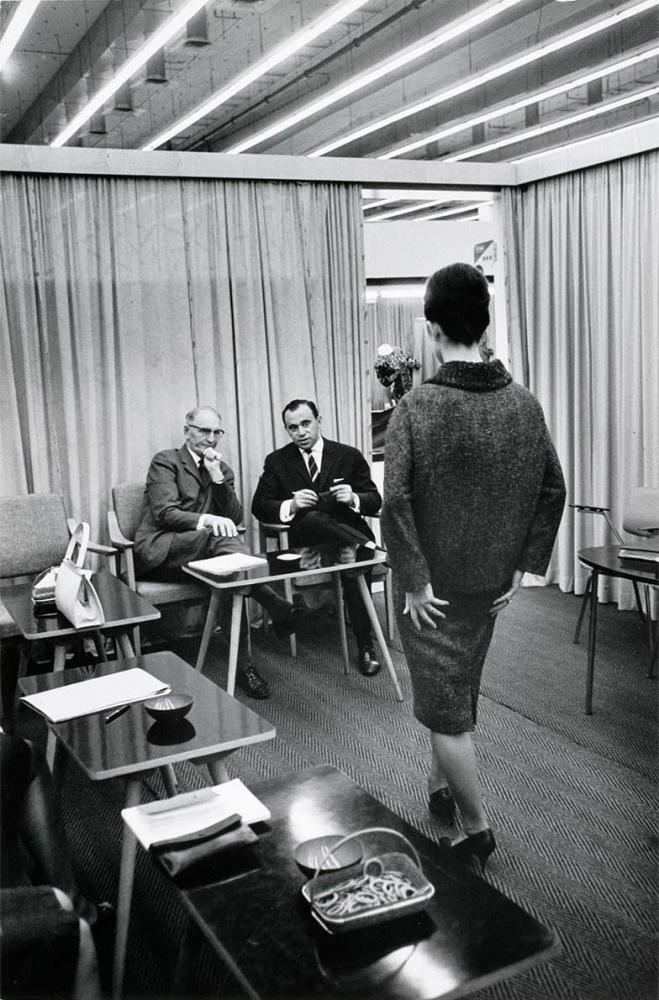 Black and white photograph: Two men in suits are sitting in front of a small table with their legs crossed, looking at a woman in a costume. The woman is standing in the foreground with her back to the viewer. Curtains and a mirror can be seen behind the men.