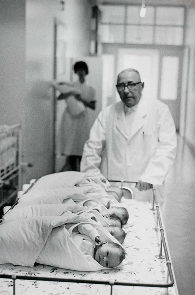 Black and white photograph: An elderly gentleman in a white doctor’s coat pushes a trolley with six newborn babies, behind him in the corridor is a nurse with a bundle in her arms.