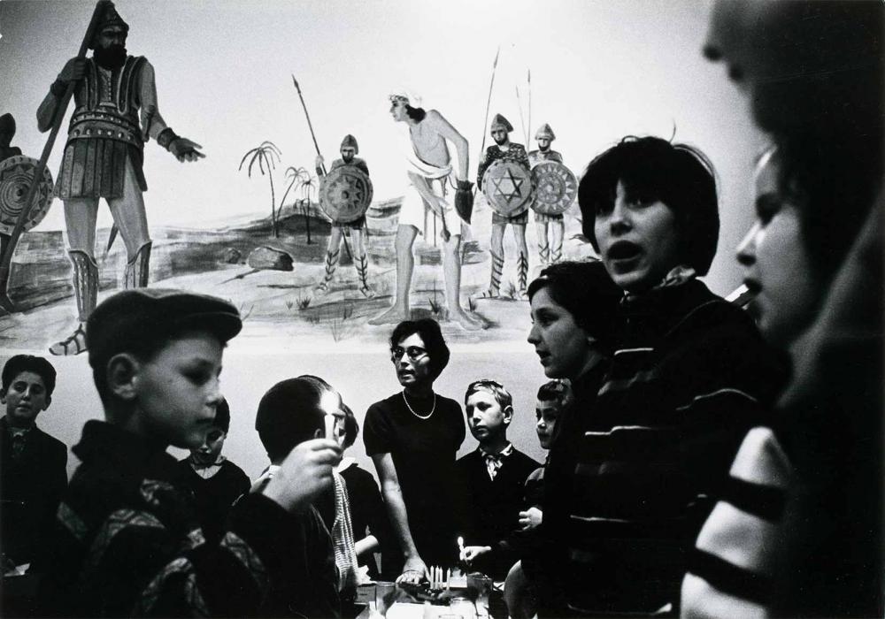 Black and white photograph: The photo shows a lady and several children, two of the children are holding a candle. They are in a room and surround a table. The wall in the background depicts the battle of David against Goliath.