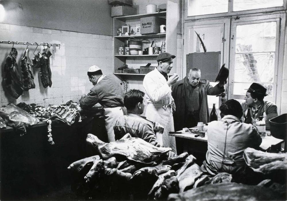 Black and white photograph: View into a slaughter room. In the right half of the picture are six men, four of whom are wearing headgear. The left half of the picture shows cut-up meat on tables and on hooks on the wall.