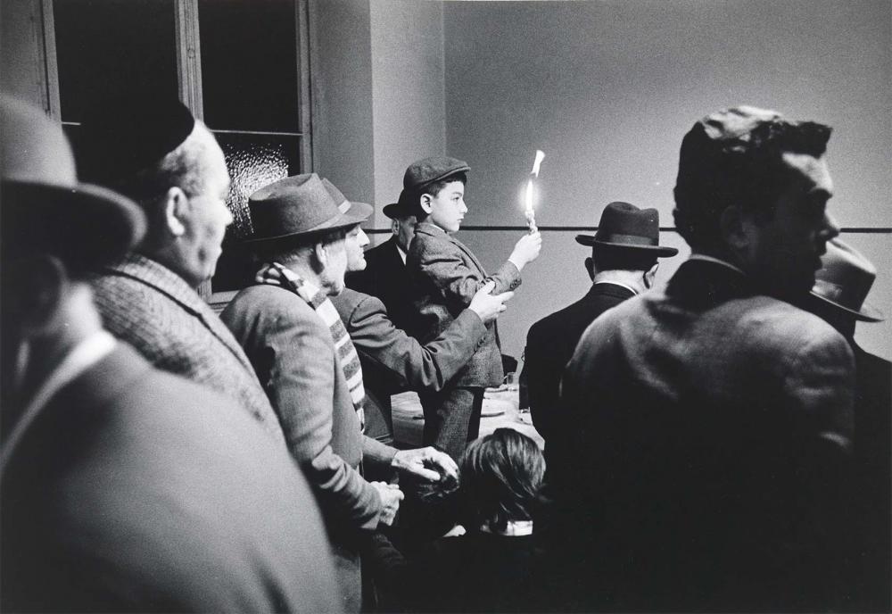 Black and white photograph: Eight men and three children can be seen in a room. All but two of the children are wearing headgear. They are standing close together in rows. One boy is standing slightly higher and holding a lit candle in his right hand. A man is holding the child’s arm for support.