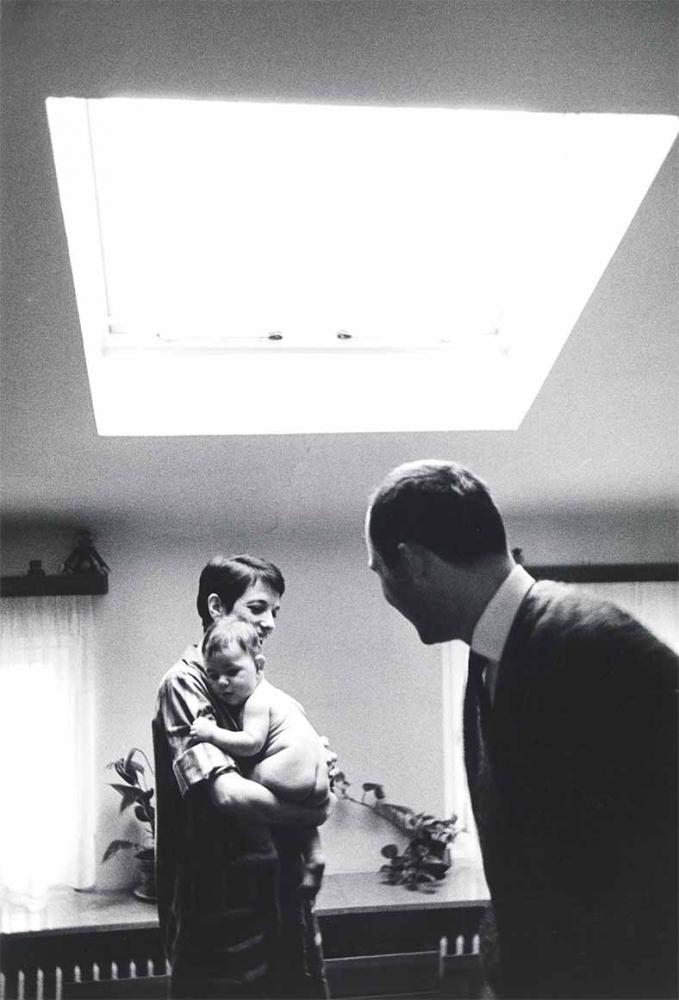 Black and white photograph: A man and a woman are standing in an apartment with a skylight. She is holding an unclothed baby in her arms. Two other windows with curtains and two potted plants on a ledge can be seen in the background.